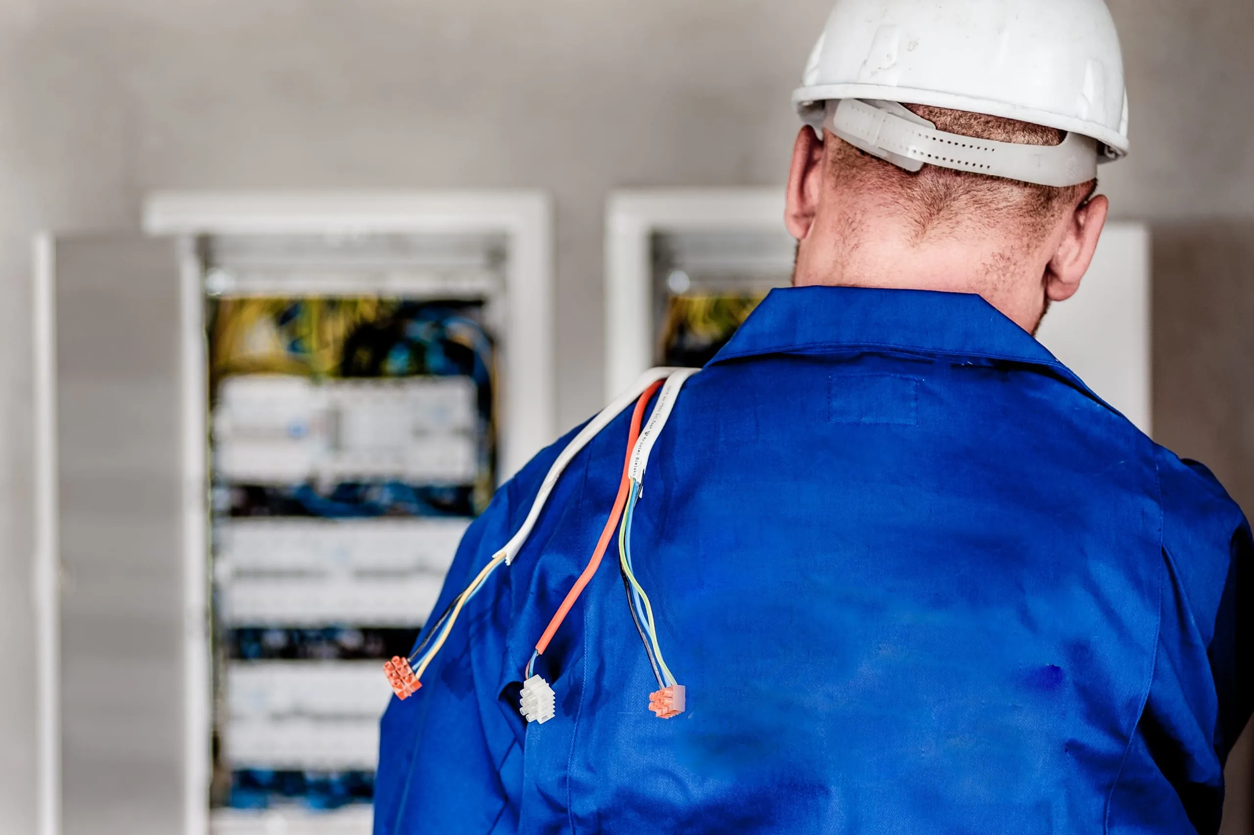 An electrician in a white hard hat and blue overalls at an open switchboard, seen from behind