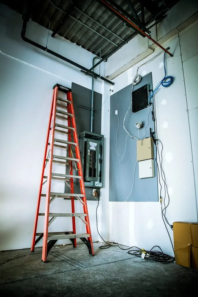 An orange ladder beside an open electrical panel on a white wall